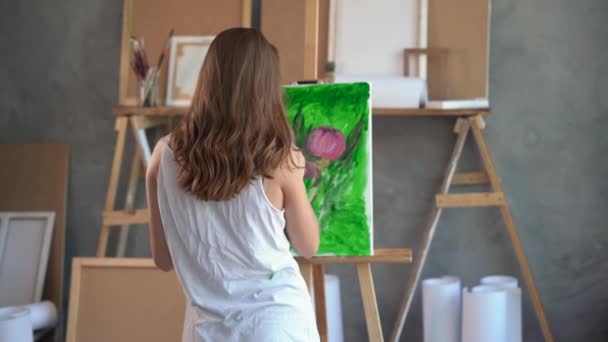 Achteraanzicht van een vrouw met haar, gekleed in een wit T-shirt gekleurd met verf, staat in een kunststudio en schildert een olieverf. Gereedschap is verspreid over. — Stockvideo