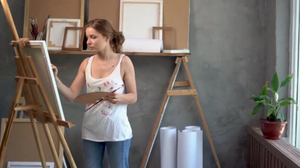 Mulher artista concentrado que trabalha em oficina de arte pinta uma obra-prima. Retrato de uma pessoa criativa com cavalete, paleta, pincel e tintas. Conceito de espaço arte. — Vídeo de Stock