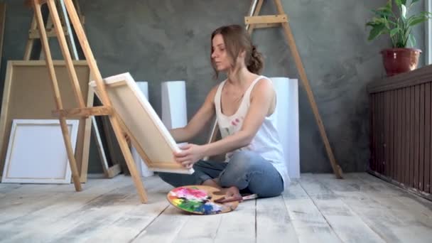 Una artista examina una pintura una obra maestra sentada en un taller creativo con un caballete y un lienzo. el artista se regocija por el éxito sostiene la imagen sonriendo. concepto de arte. — Vídeo de stock