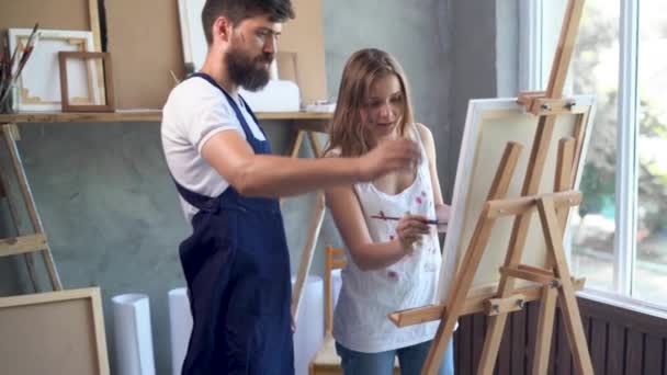 Um par de jovens artistas que trabalham em um estúdio de arte criando uma pintura em tela. O artista barbudo ensina sua namorada a pintar lona com tintas, a usar obras abstratas na oficina. — Vídeo de Stock