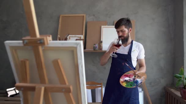 Homme barbu - un artiste peint dans un atelier d'art avec un verre de vin et une palette dans ses mains. Peintre est une crise créative réfléchie. Manque d'idées. Concept de créativité. — Video