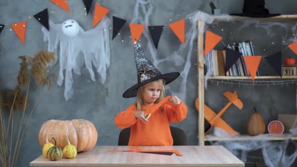 Artigianato di Halloween. Ragazza caucasica in un cappello da strega si siede a casa a tavola e forbici taglia zucche e fantasmi di carta. — Video Stock