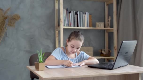 Linda adolescente colegiala haciendo la tarea con el ordenador portátil en casa. El niño escribe la tarea en un cuaderno. — Vídeos de Stock