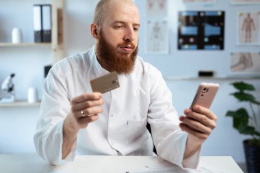 Mobile shopping. Bearded man doctor using smartphone shopping online working in hospital office holding credit card making payment. Medicine, banking and E-Commerce concept