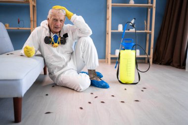 Pest control exterminator elderly man in protective suit sitting on floor near cockroaches holding head with omg troubled expression face after exterminating roaches under sofa in house