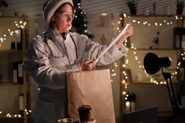Gözlüklü genç bayan doktor ve Noel Baba şapkası Noel arifesinde hastanede Noel arifesinde çalışarak paket paketini kağıt torbalara boşaltıyor. Tatil, ilaç ve tüketim