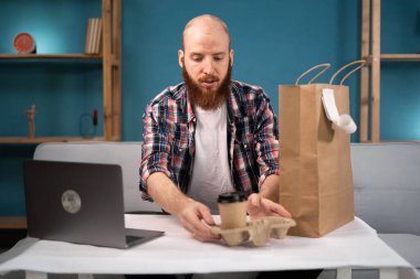 Kareli gömlekli sakallı adam evde dizüstü bilgisayarla masada oturmuş abur cubur ve kahve paketleri açıyor. Tüketim, yeme ve insanlar konsepti