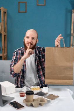 Yemek servisi, tüketim ve yeme konsepti. Kareli gömlekli sakallı bir adam, elinde paket yiyecekle sessizlik jesti yapıyor. Boşluğu kopyala