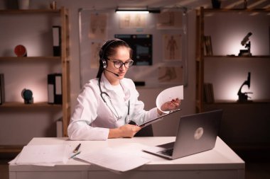 Kulaklıklı kadın doktor hastayla sohbet ediyor dizüstü bilgisayarda danışmanlık yapıyor, hastanede gece vardiyasında kulaklık ve bilgisayar kullanıyor. teletıp kavramı