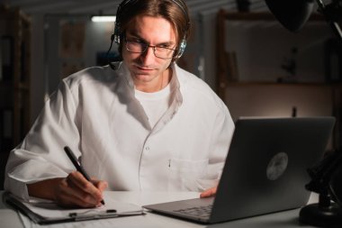 Doktor geç saatlere kadar ya da fazla mesaiye kalıp dizüstü bilgisayar kullanıyor ve notlar yazıyor. tele sağlık. Erkek doktor, hastanede gece vardiyasında çalışan sağlık bilgilerini kaydediyor. Boşluğu kopyala