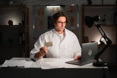 Gece vardiyası sırasında ofisindeki dizüstü bilgisayarı kullanarak online alışveriş yapan çok çalışan bir doktor. Boşluğu kopyala