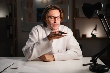 Gözlüklü ve beyaz önlüklü bir doktor elinde akıllı telefon ile hoparlörden sesli mesaj kaydediyor. Tıp, sağlık, uzaktan iletişim, ses tanıma ve yapay zeka kavramı