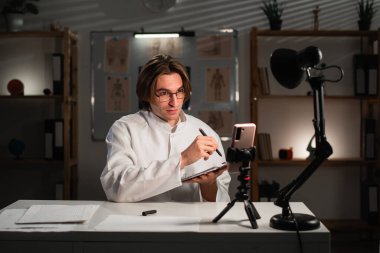 Teletıp konsepti. Erkek doktor, akıllı telefon danışmanı hastasını internette kullanıyor, hastanede gece vardiyasında çalışan notlar alıyor. Doktor terapist video görüşmesi yapıyor. Boşluğu kopyala