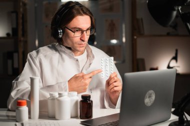 Genç erkek doktor bir ofiste gece vardiyasında reçete yazan bir hap tutuyor. Boşluğu kopyala