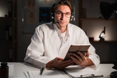 Kulaklık takan genç doktor elinde dijital tabletle klinikte gece vardiyasında çalışırken sana gülümsüyor. Boşluğu kopyala