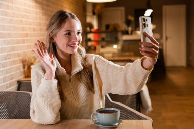 Genç, gülümseyen bir kadın cep telefonuyla video görüşmesi yapıyor. Çatı katındaki kafede kahve molası sırasında akıllı telefona el sallıyor. Boşluğu kopyala