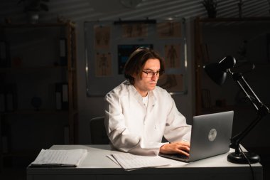 Gözlüklü genç doktor klinikteki dizüstü bilgisayarla gece vardiyasında çalışıyor. Boşluğu kopyala