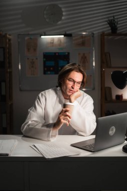 Üniformalı erkek doktor gece vardiyasında çalışıyor ve sıcak kahve içiyor. Boşluğu kopyala