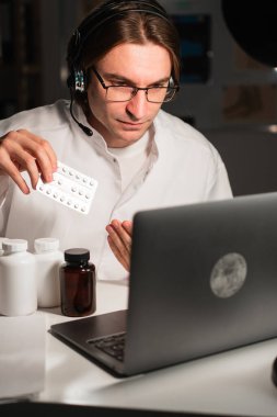 Kulaklıklı erkek doktor ve elinde ilaç şişesi olan beyaz önlüklü ve hastanede gece vardiyasında çalışan hastalara tavsiye edilen haplar. Boşluğu kopyala
