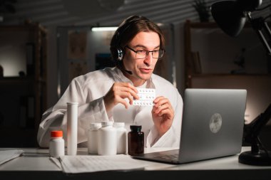 Kulaklıklı erkek doktor ve elinde ilaç şişesi olan beyaz önlüklü ve hastanede gece vardiyasında çalışan hastalara tavsiye edilen haplar. teletıp kavramı