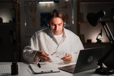 Gözlüklü bir erkek doktor, ofisinde gece vardiyasında çalışan dizüstü bilgisayarla çevrimiçi video görüşmesi yapıyor. Teletıp kavramı