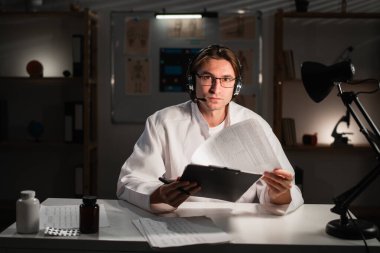 Gözlüklü bir erkek doktor, ofisinde gece vardiyasında çalışan dizüstü bilgisayarla çevrimiçi video görüşmesi yapıyor. Teletıp konsepti. Boşluğu kopyala