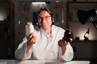 Doktor ilaç, hap ve ilaç öneriyor. Kulaklıklı ve gözlüklü genç terapist hastanede gece vardiyası sırasında kameraya bakan hap kavanozunu gösteriyor. Boşluğu kopyala