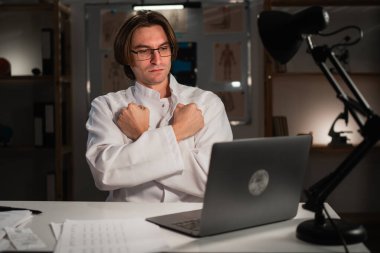 Sinirli doktor, hastanede gece vardiyası sırasında dizüstü bilgisayar kullanarak blok hareketi yaptığını gösteren video konferansı yapıyor. Hastayla çevrimiçi konuşma. Boşluğu kopyala