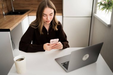 Evdeki mutfakta dizüstü bilgisayarla çalışan akıllı telefon kullanan genç bir kadın. Öğrenci internette akıllı telefon sörfü yapıyor. en üstteki görünüm