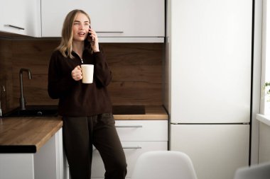 Rahatlamış bir kadın, elinde bir fincan kahve ile mutfağın içinde telefonla konuşuyor. Boşluğu kopyala