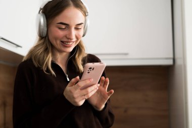 Evdeki mutfakta akıllı telefon kullanan kulaklık takan genç bir kadın. Boşluğu kopyala