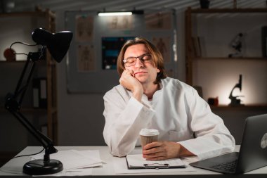 Üniformalı doktor, klinikte gece vardiyası sırasında masada otururken elleriyle poz vermekten yoruldu. Boşluğu kopyala