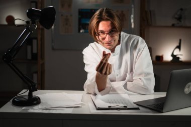 Gözlüklü pratisyen doktor hastane ofisinde gece vardiyasında çalışırken para hareketi yapıyor. Boşluğu kopyala