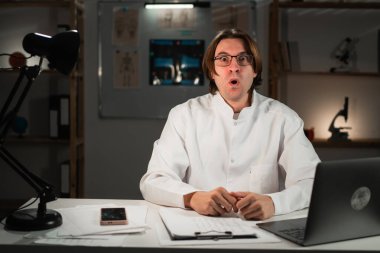 Omg. Şok olmuş pratisyen doktor gece vardiyasında çalışıyor hastane ofisindeki kameraya bakıyor. Boşluğu kopyala