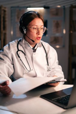 Beyaz önlüklü doktor ve kulaklıklı doktor bilgisayarla gece vardiyasında çalışan bir hastaya çevrimiçi video görüşmesi yapıyor. Teletıp kavramı