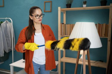 Genç bir kadın oturma odasında toz fırçasıyla lambayı temizliyor. Boşluğu kopyala