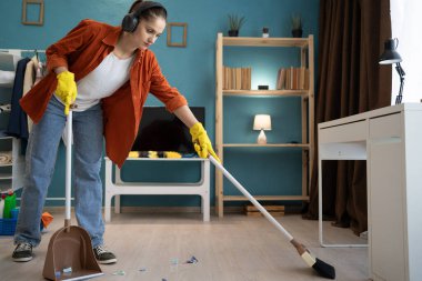 Kulaklıklı genç bir kadın süpürgeyle yerleri süpürüyor. Ev temizliği, ev işi ve temizlik konsepti. Boşluğu kopyala