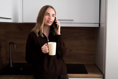Rahatlamış bir kadın, elinde bir fincan kahve ile mutfağın içinde telefonla konuşuyor. Boşluğu kopyala
