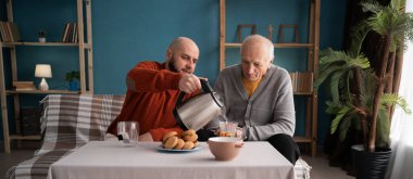 Adult man with his grandfather drinking tea or coffee sitting on sofa in the living room. family spending time concept
