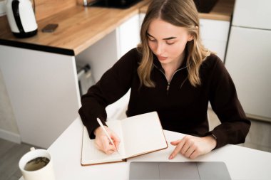 Genç bir kadın internetteki sanal giysiyi incelemek ve notlar yazmak için dizüstü bilgisayar kullanıyor. Evde çalışıyorum. Boşluğu kopyala