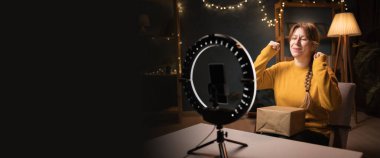 Kadın blogcu akıllı telefon kullanarak kutu kayıt videosunu açıyor. Gece geç saatlerde merkezde kutlama yapan tripodlu ışık yanıyor. İnternet alışverişi ve sosyal medya kavramı. Pankart