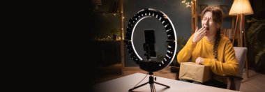 Kadın blogcu, akıllı telefon kullanarak video kaseti açıyor. Gece geç saatlerde ev ofisinde eli ağzında üç ayaklı esniyor. Çevrimiçi alışveriş ve sosyal medya kavramı