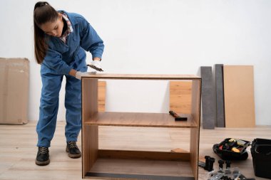 Mobilya montaj hizmeti kavramı. Yeni raflar ve dolap monte ediliyor. İş kıyafetli bir kadın ev içinde ahşap bir mobilya monte ediyor. Boşluğu kopyala