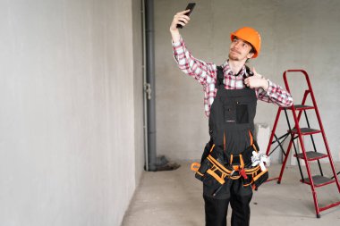 Builder, foreman, repairman wearing tool belt hard hat and overalls at construction site making point of view photo showing thumb up, like gesture. Banner. copy space