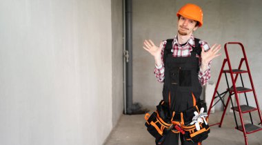 İnşaatçı, ustabaşı, alet kemeri takan bir tamirci inşaat alanında kol ve ellerle çaresiz bir hareket sergiliyor. Pankart. Boşluğu kopyala