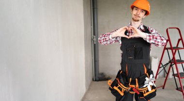 İnşaatçı, ustabaşı, alet kemeri takan tamirci inşaat alanında kameraya aşk jesti gösteriyor. Pankart. Boşluğu kopyala