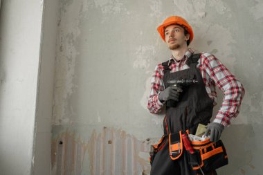 Duvarda delgi tutan genç işçi ya da tamirat sırasında arka planda elektrikli matkap. Boşluğu kopyala