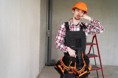 Erkek işçi, inşaatçı ya da tamirci telefon jesti yaparak beni arayın diyor, tulum, alet kemeri ve koruyucu başlık takıyor, inşaat alanında çalışıyor. Boşluğu kopyala