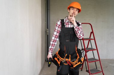 Erkek işçi, inşaatçı ya da tamirci sessizlik işareti göstererek tamirat ya da bina onarımına hazır, tulum, alet kemeri ve koruyucu kask giyiyor, inşaat alanında çalışıyor. Boşluğu kopyala