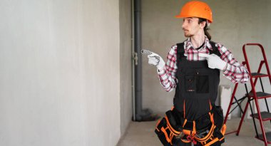 Erkek işçi, müteahhit ya da tamirci, inşaat noktası işaret parmağını bölgeye doğru kaldırmaya hazır, tulum, alet kemeri ve koruyucu kask takıyor, inşaat alanında çalışıyor. Boşluğu kopyala
